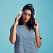 © peopleimages.com - Portrait, headphones and Indian woman streaming music, audio and listening to sound on blue studio background. Serious face, person and girl with headset and gen z with attitude and app for radio