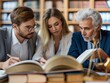 © MaxK - Three people are sitting at a table with books and papers in front of them. They are looking at a book and writing in a notebook. Scene is focused and studious