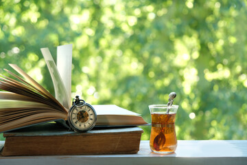 Wall Mural - Glass tea cup, pocket watch and old books on wooden table in garden, green natural background. spring, summer season. atmosphere gentle image. Harmony, relax atmosphere