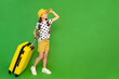 A little girl with a suitcase on wheels looks into the distance. A child in shorts and a hat is eating a vacation at the sea. Green isolated background. Copy space.