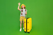 A beautiful little girl is getting ready to travel with a big yellow suitcase. A child in shorts and a hat is eating for the summer holidays and waving his palm. Green isolated background.