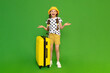 A little girl with a big suitcase on wheels, wearing a hat and shorts. Summer holidays for children.  The child spread her palms in surprise and smiles broadly. Green isolated background.