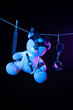 A toy teddy bear in a bdsm mask and leather straps hangs on a clothesline in neon light on a dark background
