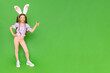A little girl with rabbit ears on her head points to your ad. The child is standing in full height dressed as an Easter bunny, enjoying the holiday. Green isolated background. Banner. Copy Space