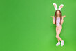 A little girl with rabbit ears on her head points to your ad. The child is standing in full height dressed as an Easter bunny, enjoying the holiday. Green isolated background. Banner. Copy Space