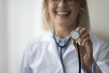 Fototapeta Tulipany - Close up of smiling mature woman cardiologist showing to camera stethoscope. Assessing patient health, listen internal body sounds, check health condition of heart or lungs, measure of blood pressure