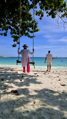 Wall Mural - A man and a woman are enjoying the view of the azure sky and water while standing next to a swing on the sandy beach, surrounded by palm trees, Koh Samet Thailand