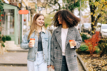 Two happy carefree multiracial girlfriends walking city street with take away coffee, talking and sharing life stories, multiethnic female best friends