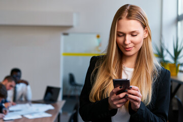 Professional blonde woman in smart casual attire uses smartphone in modern office. Focused businesswoman engages with mobile tech, managing tasks. Colleagues collaborate in background workspace.