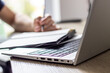 © Point of view - a man makes notes in a notebook ,typing in laptop ,at a table, image in light colors, online lesson or online meeting, cafe or office, modern silver laptop, blurred window background