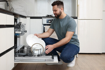 Canvas Print - Smiling man loading dishwasher with plates in kitchen