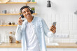 Happy positive African American man dancing and listening to music on headphones at home kitchen