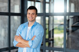 Fototapeta Kuchnia - Young successful Caucasian man entrepreneur or an office worker stands with crossed arms in a modern office, looking at the camera and smiling