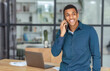 Young successful African American man entrepreneur or an office worker stands in a modern office and talking on a mobile phone, smiles friendly