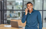 Fototapeta Kuchnia - Young successful African American man entrepreneur or an office worker stands in a modern office and talking on a mobile phone, smiles friendly