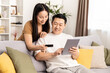 © puhhha - Happy Couple Shopping Online with Credit Card at Home. Cheerful man and woman sitting on sofa, shopping on tablet with credit card in a cozy living room.