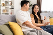 Smiling Asian couple sitting on a couch, engaging in a light-hearted conversation while enjoying a warm cup of coffee in a cozy living room.