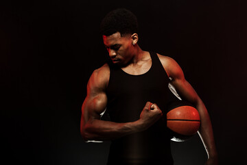 Wall Mural - Basketball player side lit with red color holding a ball against black background. Serious muscular african american man showing muscles