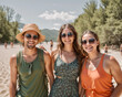 © stopcontrol - Three smiling friends with sunglasses enjoying a warm, sunny day on a sandy beach with other beachgoers in the background.