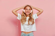 photo of happy blonde girl wearing headphones, standing against pastel pink background, white tshirt