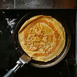 
A top-down shot of a crepe being prepared on a hot griddle, showing the batter being spread thinly and cooked until golden brown, with a spatula ready to flip it.