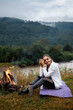 Travel, tourism, camping. Young calm woman tourist at the beautiful nature landscape sitting near the tent by the fire. Girl with drink in mug in her hands