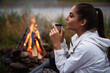 Side view of caucasian female tourist drinking hot tea while sitting near bonfire. Pretty woman enjoying amazing view on river and green nature during camping.