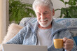 Senior bearded smiling man sitting on sofa looking at laptop holding a coffee cup - indoor, at home concept - caucasian elderly retired man using technology