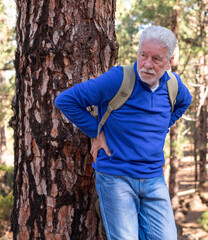 Sticker - Senior white-haired man hiking in the mountains leans against a tree trunk to catch his breath, touching his back. The elderly pensioner is not feeling very well