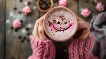 Pink Cup Of Coffee With Pieces Of Chocolate And A Smiley Face On The Foam In Hands In A Pink Knitted Sweater On A Wooden Table In A Cozy Atmosphere. The Concept Of Coziness, Breakfast, Chocolate Day