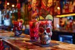 In a festive setting, two skull-shaped glasses adorned with a chili powder rim are filled with vibrant red cocktails, garnished with lime wheels and maraschino cherries.