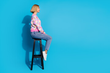 Wall Mural - Full length side profile photo of positive girl wear pullover sit on bar chair look at offer empty space isolated on blue color background