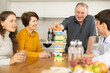 Cheerful senior man spending free time with wife and adult children at laid-back family home get-together with drinks, concentrating on removing block from tower while playing jenga