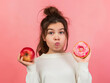 Uma jovem em fundo rosa, segurando uma rosquinha em uma mão e uma maçã na outra, refletindo sobre nutrição, saúde dieta e escolhas alimentares