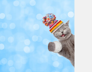 Aufkleber - Happy cat wearing warm winter knitted woolen hat with pompon  holds snowball behind empty white banner. Blurred blue background