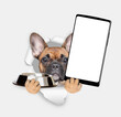 French bulldog puppy looks through a hole in white paper, holds empty bowl and  shows smartphone with white blank screen in it paw, Empty free space for mock up, banner