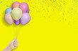 Female hand holds bunch of colorful balloons on yellow background with confetti. Empty space for text