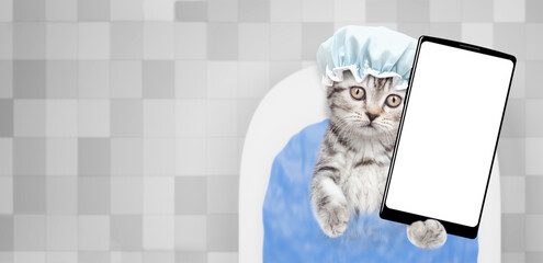 Fototapete - Cute kitten wearing shower cap takes the bath at home and shows big smartphone with white blank screen in it paw. Top down view. Empty space for text