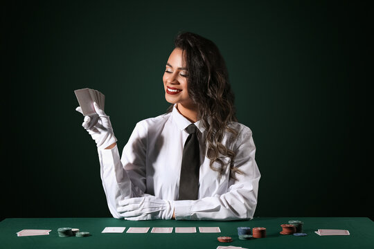 Beautiful African-American woman sitting at poker table with playing cards and chips on dark green background