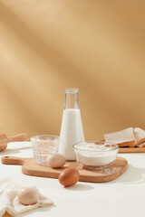 Bowl of flour, eggs and milk bottle featured on wooden chopping board against milk coffee brown background on white kitchen counter. Front view photo with copy space