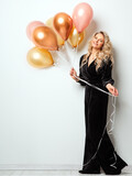 Fototapeta  - Beautiful Woman with Balloons over White background. Birthday Party Time. Fashion Model with Curly Hairstyle in Black Long Dress