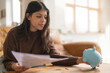 © Iryna - confident young indian woman holding piggi bank and letter invoice savings employment tax paying energy bill ready to use savings