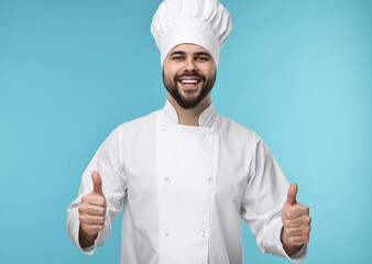 Wall Mural - Happy young chef in uniform showing thumbs up on light blue background