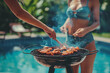 Lifestyle photo of friends having an outdoor barbecue in the backyard with a pool party background. A woman is grilling meat on a grill barbecue at home during summer vacation.