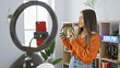 © Krakenimages.com - A young hispanic woman records a video indoors using her phone with a ring light, portraying a casual and attractive music studio vibe.