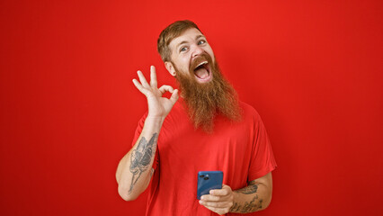 Wall Mural - Beaming redhead guy giving ok sign while engrossed in messaging on his smartphone, standing confidently against a vibrant red wall, exuding joy, and positivity.