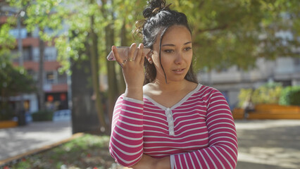 Poster - A young hispanic woman listens to a voice message on her phone while standing outdoors in a city park.