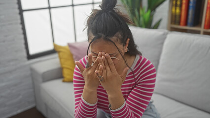 Wall Mural - A young, distressed hispanic woman covering her face with hands, sitting on a white sofa in a bright, modern indoor setting.