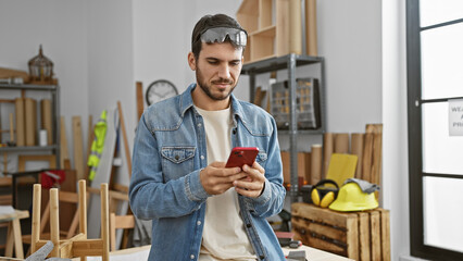 A young hispanic man in casual attire uses a smartphone in a well-organized carpentry workshop.