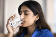 Beautiful young 20s Indian girl with closed eyes enjoying fresh pure water, drinking cold beverage from glass, satisfying thirst, keeping hydration balance, smooth healthy facial skin